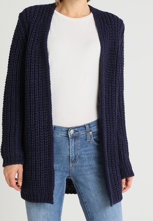 Cardigan en maille épaisse bleu marine foncé à ouverture devant porté sur une chemise blanche et un jean taille haute bleu clair.
