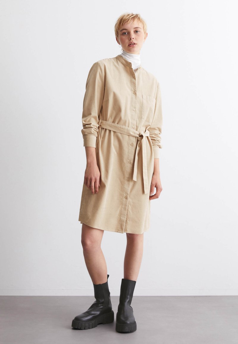 Beiges Hemdkleid aus Baumwolle mit Knopfverschluss, langen Ärmeln, einem Gürtel in der Taille und einer Brusttasche. Kombiniert mit schwarzen Stiefeletten.