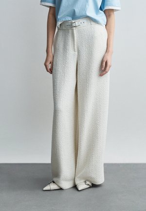 Persona vistiendo pantalones blancos texturizados de pierna ancha con cinturón a juego, camisa de manga corta azul claro, y zapatos blancos puntiagudos con hebillas.