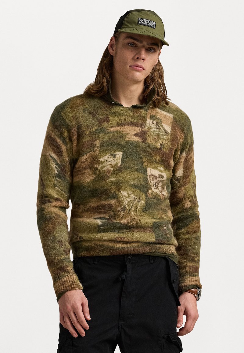 Gebreide trui in een groen en bruin camouflagedessin, met een kraag en ribgebreide manchetten. Draagt een zwarte cargo broek en een groene pet.