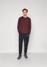 Egy férfi áll, kezét a zsebében tartva, bordó hosszú ujjú Nike pólót, fekete Nike nadrágot és színes futócipőt visel.