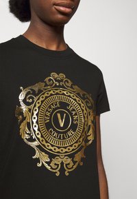 Černé bavlněné tričko s velkým zlatým potiskem loga s okrasnými detaily, které uvádí "Versace Jeans Couture" kolem centrálního emblemu.