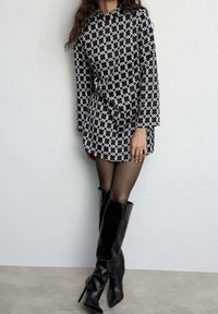 Žena v dlouhých rukávech mini šatech s černobílým vzorem, v průhledných černých punčochách a vysokých kožených botách, stojí opřená o zeď.