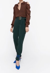 Hnědý, dlouhý rukáv, top s hlubokým V-výstřihem a volánovými rameny, spárovaný se zelenými kalhotami s vysokým pasem a cargo kapsami a modrými podpatky.