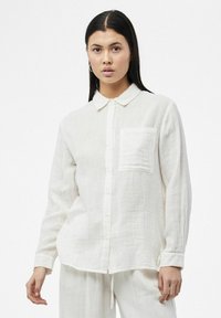 Camisa blanca de manga larga con botones, fabricada en tela texturizada, con collar y un bolsillo en el pecho. Presenta un corte relajado y un dobladillo curvado.