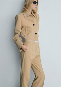 Giacca cropped beige con due bottoni neri e tasche laterali, abbinata a pantaloni beige su misura, tessuto morbido, design minimal.