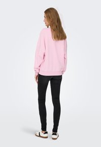 Sudadera rosa de gran tamaño con mangas largas, puños y dobladillo de canalé, combinada con jeans negros ajustados y zapatillas blancas con acentos negros.