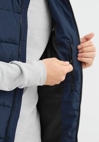 Mornarska puffer prsluk s prednjim patentnim zatvaračem; unutrašnjost ima mali džep sa patentnim zatvaračem; nosi se preko svijetlosivog majice dugih rukava.