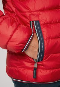 Giacca a vento rossa con cuciture orizzontali, dotata di una tasca con zip e accenti blu navy sul colletto e sull'orlo. Tessuto a trama.
