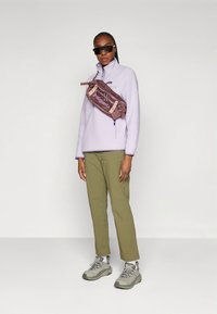 Šviesiai violetinis flisinis megztinis, alyvuogių žalių spalvų kelnės ir pilki sportbačiai. Ruda per petį nešiojama rankinė su logotipu. Modelis dėvi juodus saulės akinius.