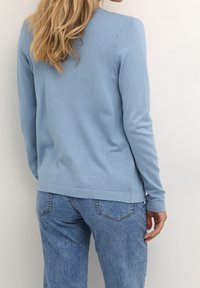 Donna che indossa un maglione a maniche lunghe blu chiaro e jeans blu di media tonalità, in piedi con la schiena rivolta verso la fotocamera davanti a un muro semplice.