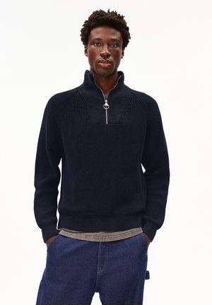 Man wearing a black knit sweater with a silver half-zip collar and blue denim pants, standing with hands in pockets, neutral background.