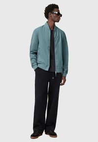 Giacca bomber verde acqua, maglietta nera, pantaloni neri larghi e scarpe nere con suole marroni; indossa occhiali da sole scuri; texture liscia.