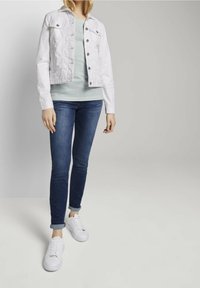 Weiße Jeansjacke mit Knopfverschluss, kombiniert mit einem grün-weiß gestreiften Oberteil, blauen Skinny-Jeans und weißen Sneakers.