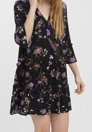 Femme portant une robe noire à fleurs violettes et roses, manches trois-quarts, et une jupe à volants, debout devant un fond uni.