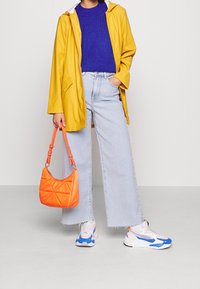 Gul vattentät jacka, blå stickad tröja, vida ljusa jeans, orange väska och vita sneakers med blå accent. Rena linjer och modern design.
