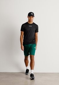 Czarna sportowa koszulka i zielone szorty, obie z logo. Model nosi czapkę w kolorze czarnym i czarne sneakersy z białymi skarpetkami, stojąc na neutralnym tle.