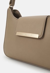 Sac à bandoulière en cuir taupe avec fermoir métallique doré embossé "ANNA FIELD" et une sangle réglable attachée par des anneaux dorés.