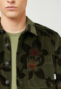 Camicia in corduroy verde con motivo floreale in marrone scuro e arancione, con chiusura a bottoni davanti e dettagli di tasche applicate.