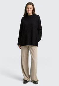 Vrouw die lacht en een losse zwarte trui, beige wijde broekspijpen en zwarte schoenen draagt, staand tegen een lichtgrijze achtergrond.