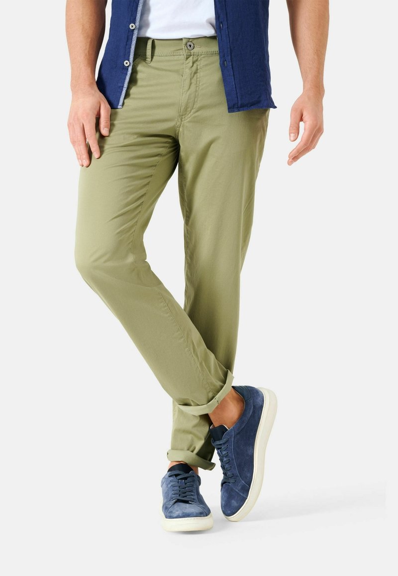 Homme portant un pantalon olive retroussé, des baskets bleu marine, des chaussettes blanches et une chemise bleue partiellement visible.