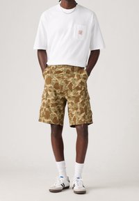 Pantalones cortos de camuflaje en beige y marrón, con dos bolsillos laterales y cierre de botón en la parte delantera. Combinados con una camiseta blanca básica y calcetines blancos.