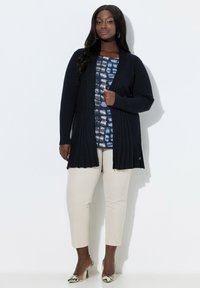 Kvinde iført en lang sort ribbet cardigan over en blå mønstret top, beige bukser og højhælede sko med slangeskindsmønster, stående mod en hvid væg.