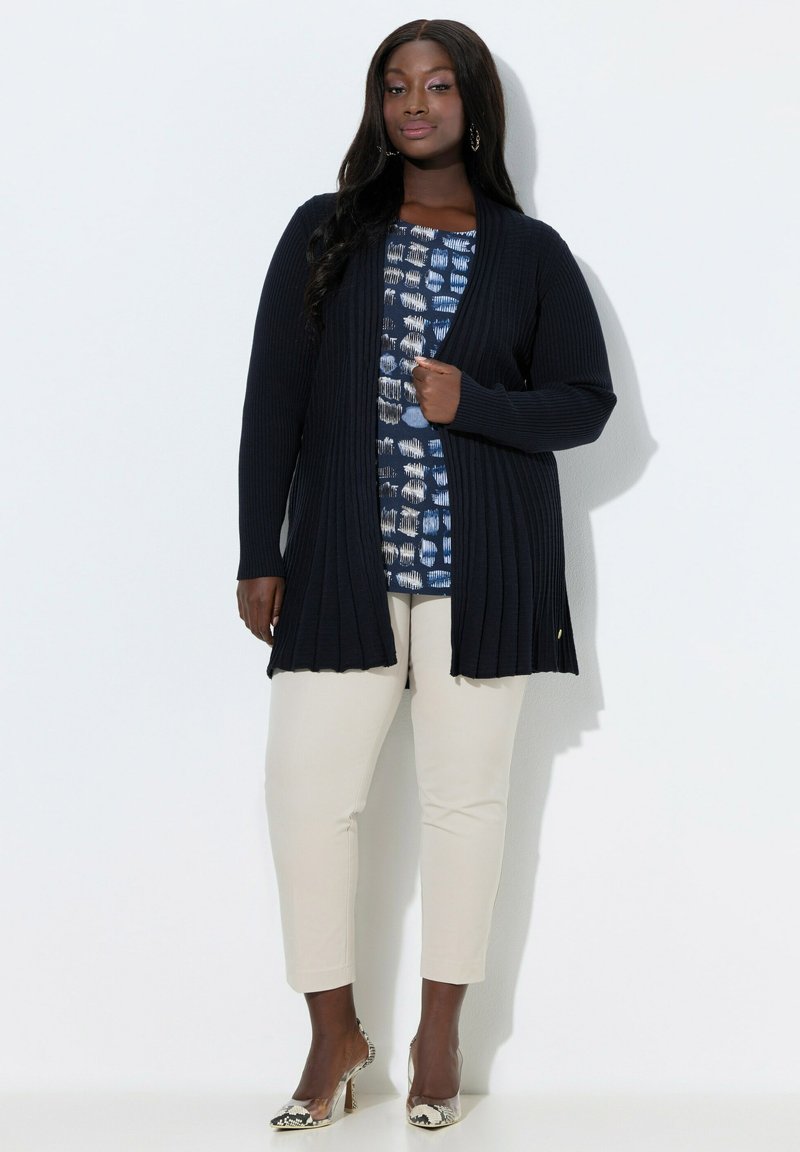 Kvinde iført en lang sort ribbet cardigan over en blå mønstret top, beige bukser og højhælede sko med slangeskindsmønster, stående mod en hvid væg.