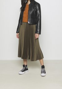Donna con giacca di pelle nera, maglione arancione, gonna midi plissettata verde oliva e sneakers alte nere in piedi su un pavimento chiaro.