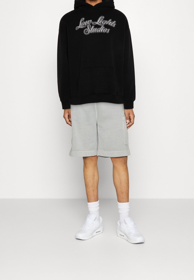 Sweat à capuche noir avec un graphisme "Low Lights Studios", associé à un short gris clair. Une paire de baskets blanches et des chaussettes courtes complètent la tenue.