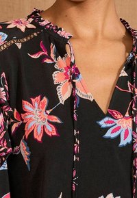 Blusa nera a fiori con fiori rosa e blu, scollo a V, colletto arricciato e delicati dettagli in pizzo lungo le maniche. Tessuto morbido.