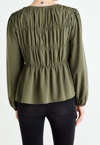 Blouse à manches longues vert olive avec texture froncée à l'arrière, dotée d'un col rond et d'un ourlet élastique. Associée à un jean noir.