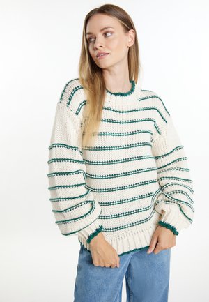 Woman wearing a white chunky knit sweater with green horizontal stripes and blue jeans, looking to the side against a white background.