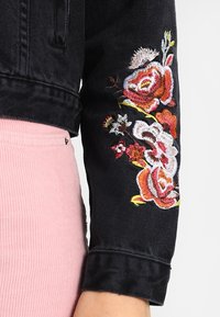 Black denim jacket featuring colorful floral embroidery on the left arm. The jacket has a cropped design and a textured fabric.