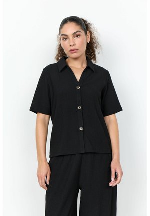 Femme aux cheveux bouclés attachés, portant une chemise noire à manches courtes boutonnée et un pantalon noir assorti, debout devant un fond blanc.