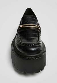 Mocassins noirs en cuir verni à texture crocodile, avec un accent métallique doré et une semelle plate-forme épaisse noire.