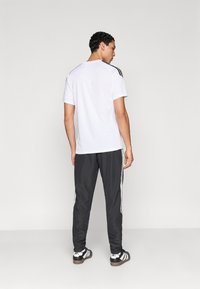 Λευκό αθλητικό κοντομάνικο με μαύρες ρίγες στους ώμους, συνδυασμένο με σκούρο γκρι παντελόνι φόρμας με λευκές πλαϊνές ρίγες. Μαύρα αθλητικά παπούτσια.