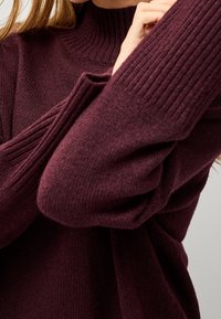 Pull en maille bordeaux à col montant avec texture côtelée, doté de larges manches qui tombent en douceur. Le matériau semble doux et chaud.