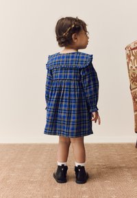 Robe à carreaux bleue avec col à volants, manches longues et taille froncée. Bottines noires avec chaussettes blanches. Vue de dos de l'enfant.