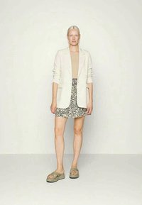 Blazer bege claro sobre um top bege, combinado com uma saia preta com estampado floral e sandálias verdes. Textura suave e corte ajustado visíveis.