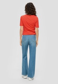 Czerwona koszulka z krótkim rękawem połączona z jasnoniebieskimi dżinsami z rozszerzanymi nogawkami. Dżinsy mają klasyczny pięciopocketowy krój i surowy brzeg. Białe trampki.