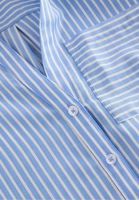 Lichtblauw overhemd met fijne witte strepen, een borstzak en een gladde stoftextuur. Vier knopen zichtbaar langs de knopenlijst.