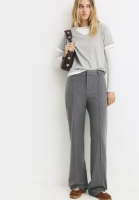 Junge Frau mit langen blonden Haaren, die gestufte graue Oberteile, hocherträgige graue Hosen, braune Sneakers trägt und eine dunkelbraune Umhängetasche hält.