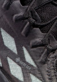 Scarpa da trekking nera con una parte superiore testurizzata, caratterizzata da dettagli riflettenti e un motivo geometrico distintivo. Design leggero e robusto.