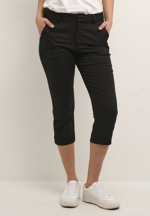 Persoon draagt zwarte cropped broek met voorzakken en witte sneakers, staand met handen in de zakken tegen een effen achtergrond.
