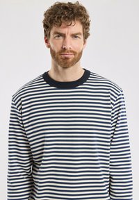 Mann mit lockigem braunem Haar und kurzem Bart, der ein navyblaues und weiß gestreiftes Langarmshirt trägt und aufrecht vor einem schlichten Hintergrund steht.