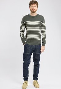 Maglione a righe verde oliva con righe crema, scollatura a coste. Pantaloni in denim blu con una vestibilità ampia e dettagli di cucitura visibili. Scarpe beige.