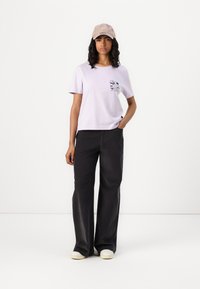 Light lavender short-sleeve t-shirt with a pocket graphic, paired with high-waisted black wide-leg pants and beige cap, set against a white background.