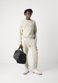 Sudadera de color beige claro con el logo "BOSS" en relieve, pantalones de chándal beige a juego con bolsillos, zapatillas blancas y un bolso de viaje de cuero negro.