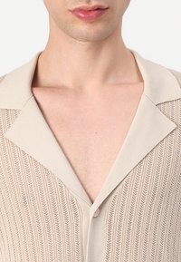 Beżowy sweter z dzianiny o fakturowanym, prążkowanym wzorze, z szerokim kołnierzem i pojedynczym zapięciem na guziki przy szyi.